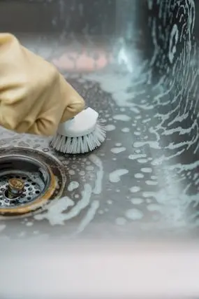 Hand in rubber glove using brush to scrub stainless steel sink with soap lather