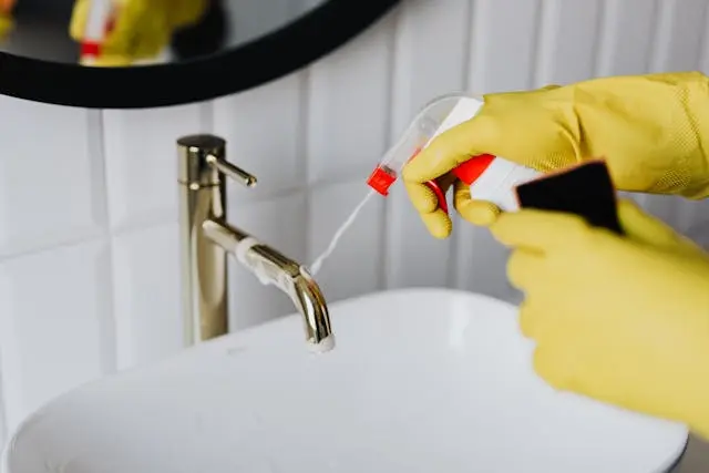 Person wearing yellow gloves cleaning bathroom faucet with disinfectant spray